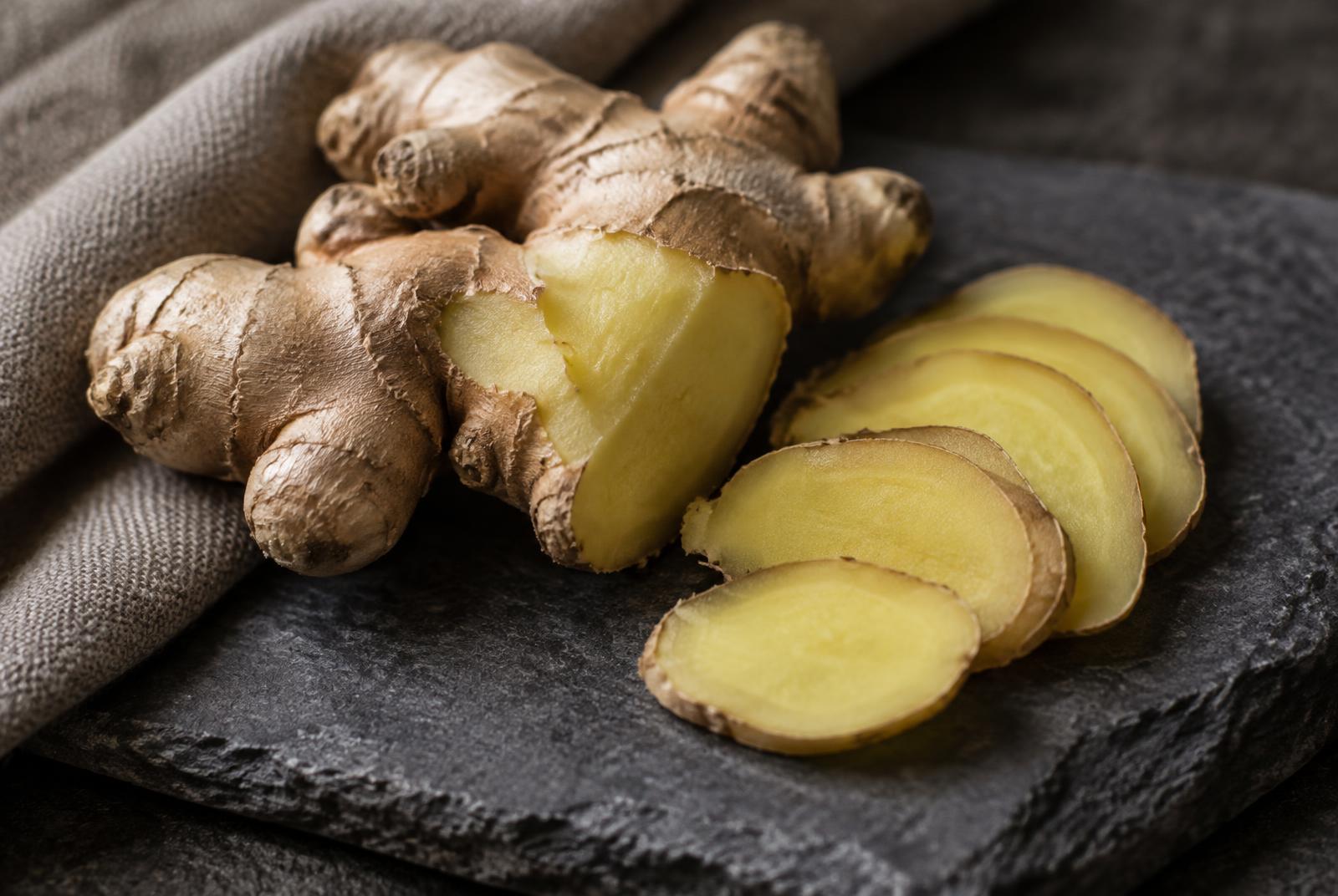 A knobbly fresh ginger rhizome cut open to show pale yellow flesh, with thin slices fanned beside it on dark stone.