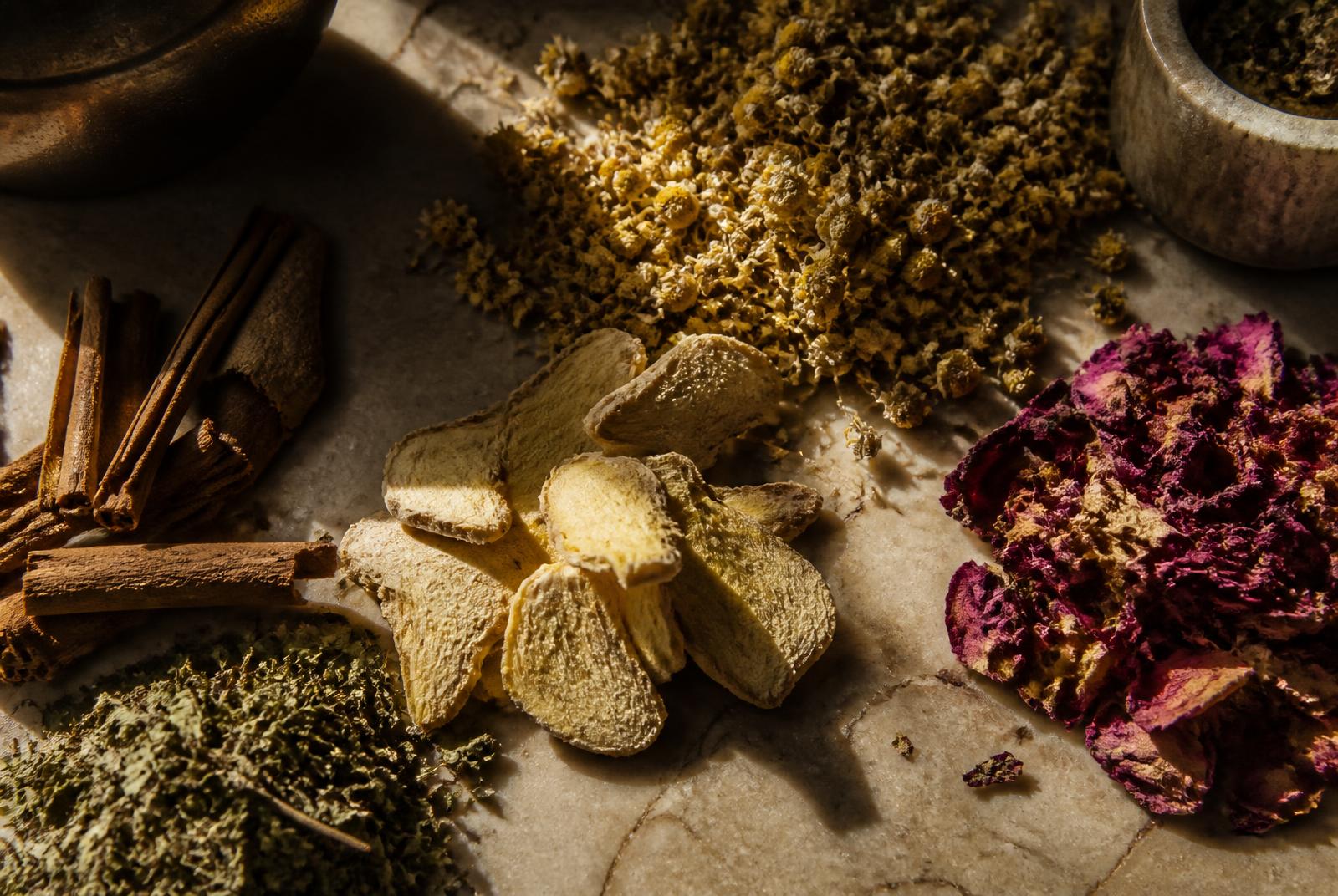 Dried botanical specimens laid out on stone: chamomile flower heads, sliced ginger rhizome, cinnamon bark, dried rose petals and loose leaf.